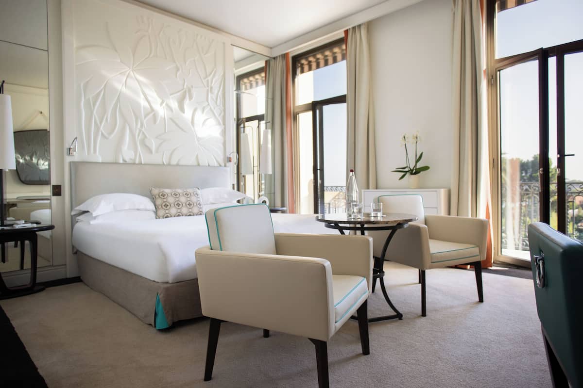 A hotel room with a bed, two armchairs, a small table, and a view of the city.