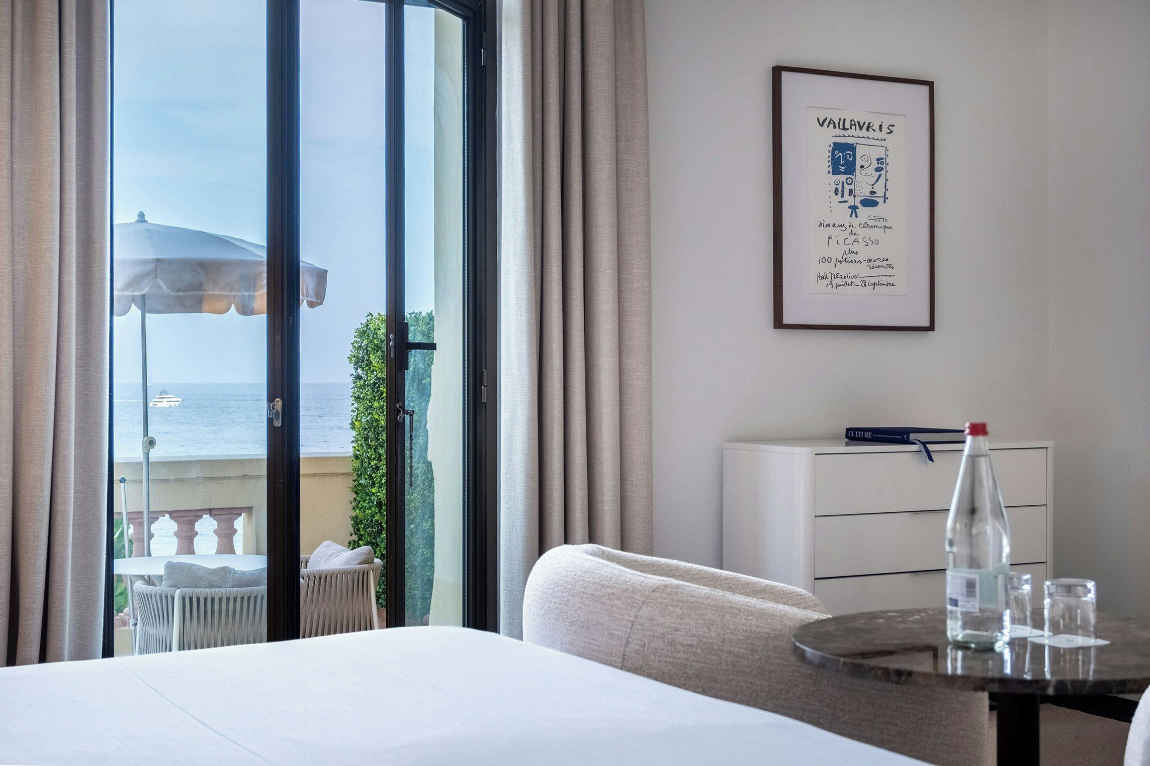 A hotel room with a bed, a table with glasses and a bottle, a framed artwork on the wall, and a view of the sea through the window.