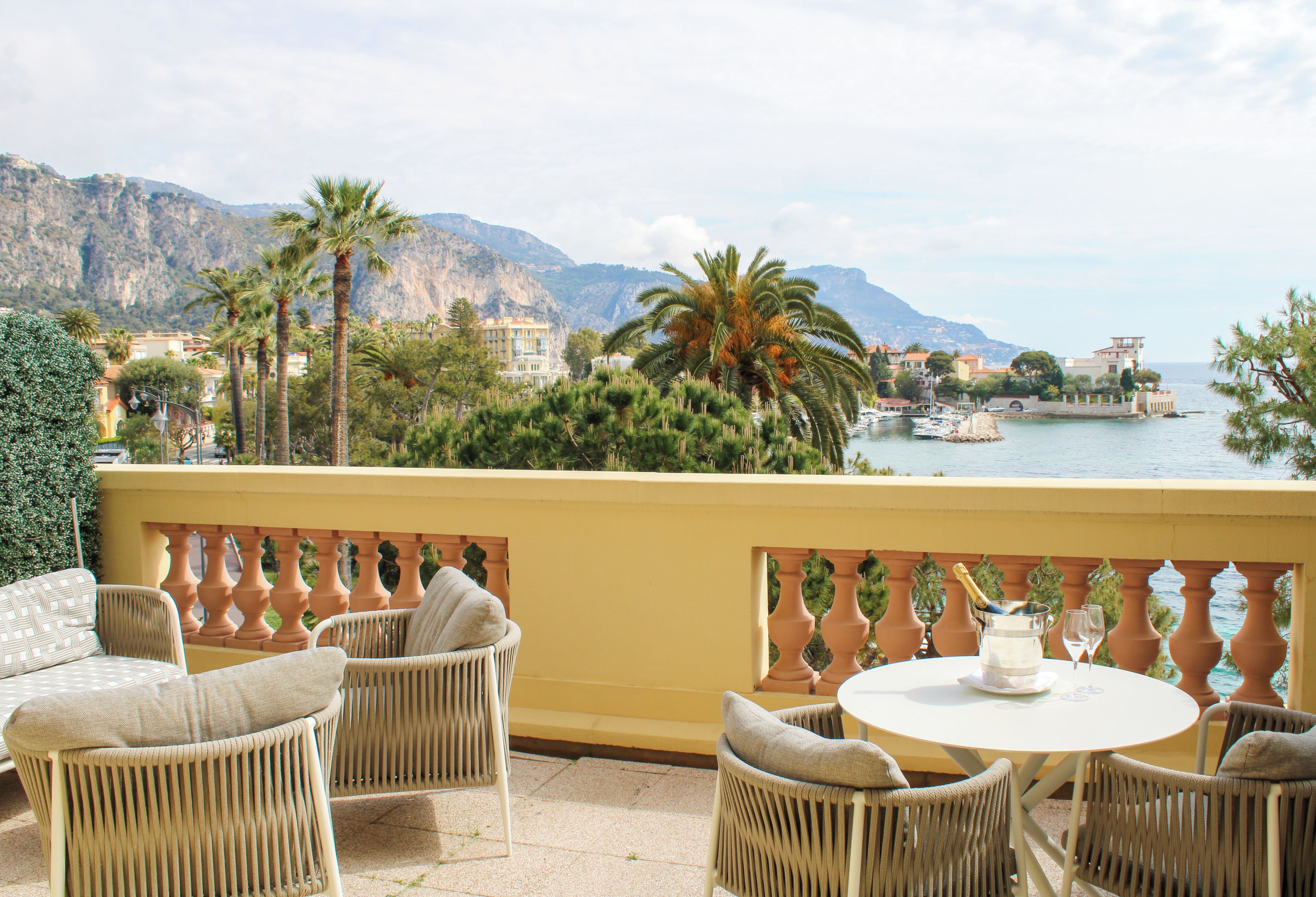 A balcony with wicker furniture, a table with a bottle and glass, and a view of a coastal town with mountains in the background.