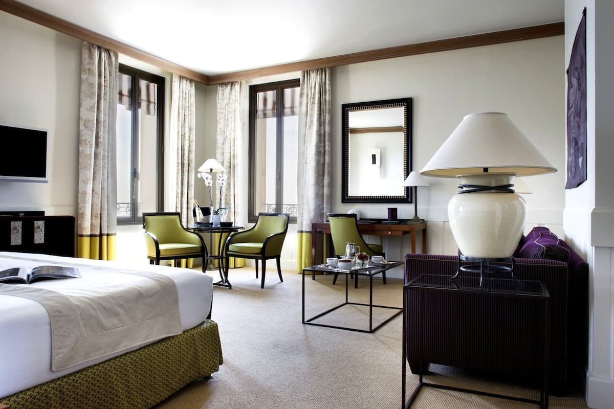 A hotel room with a bed, two chairs, a table with a lamp, a TV, and a large window with curtains.
