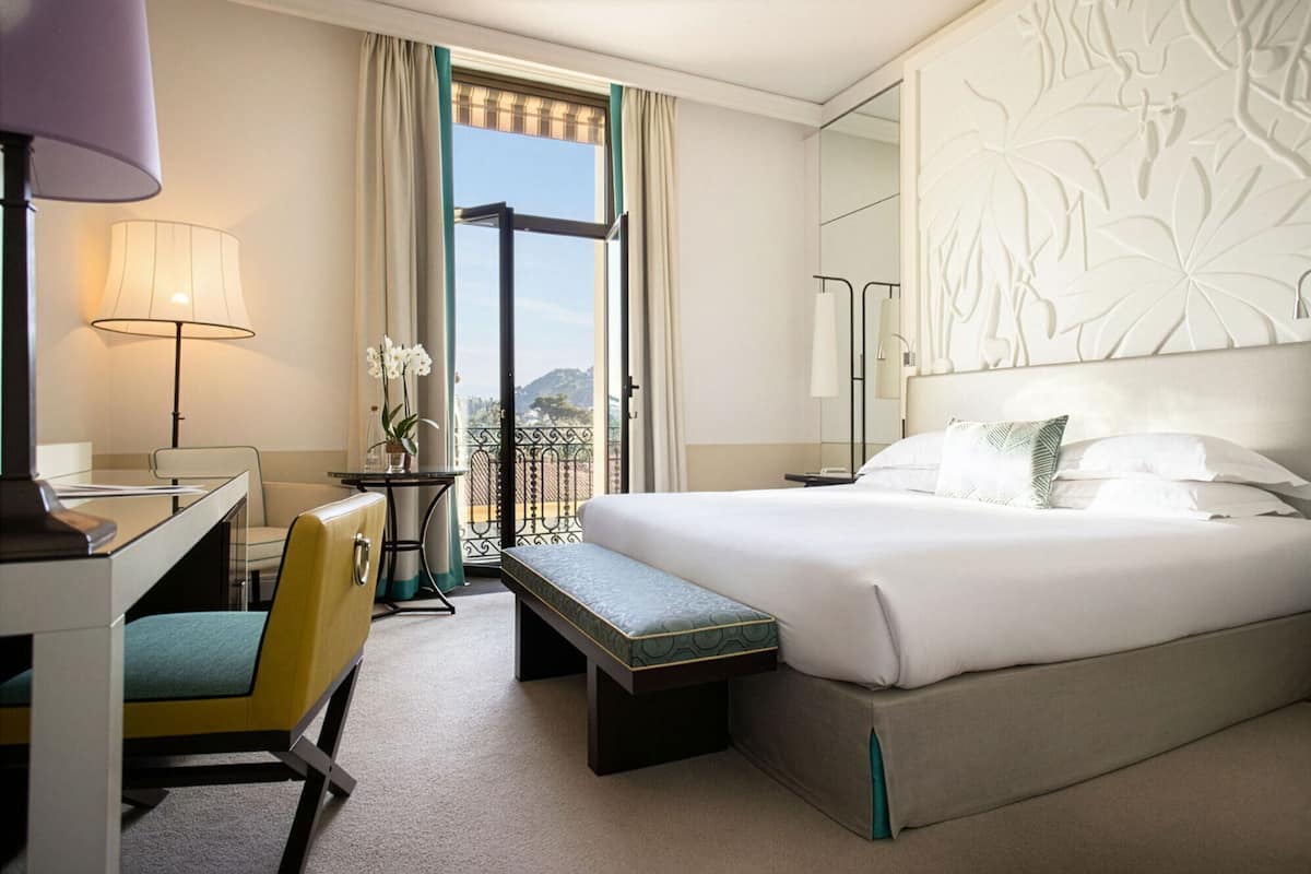A hotel room with a bed, desk, chair, lamp, and a view of the city through a window.