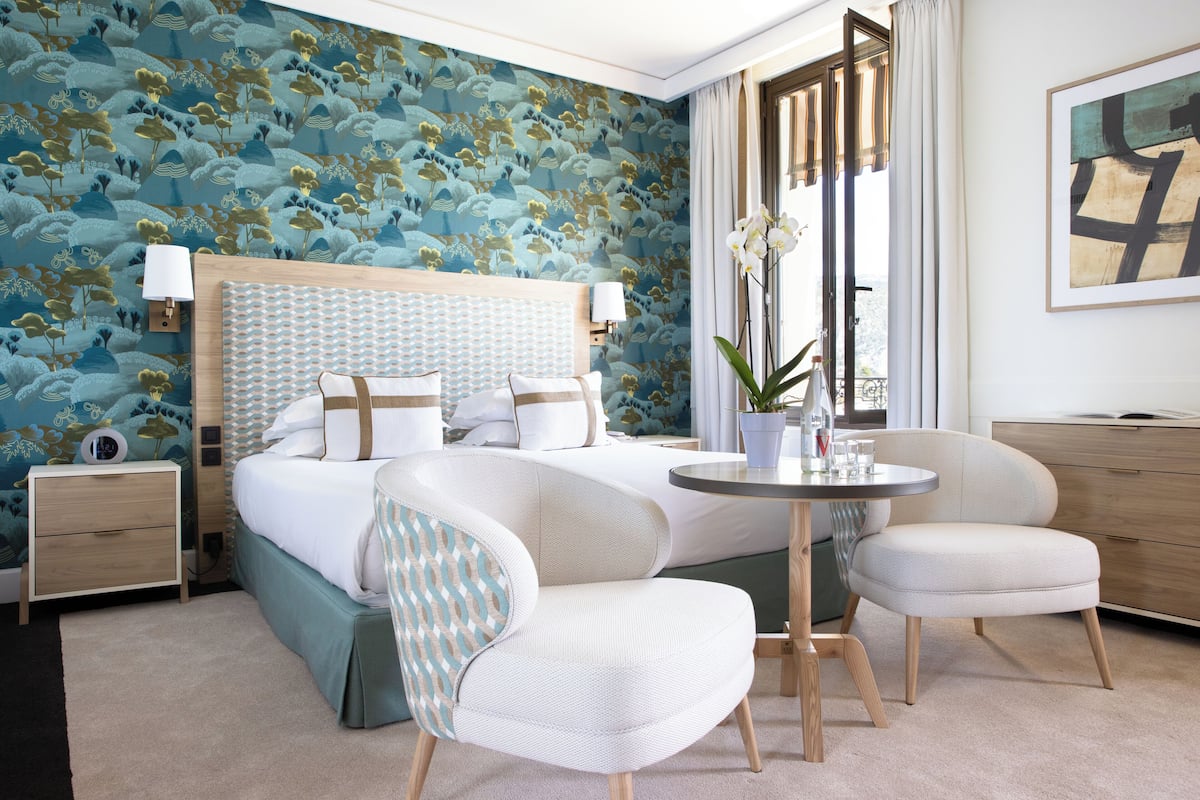 A hotel room with a large bed, two chairs, a small table, a nightstand, and a window with curtains.