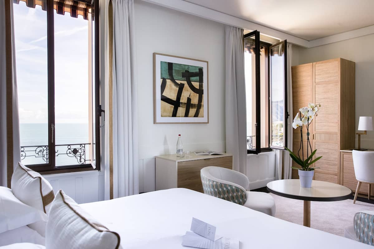 A hotel room with a bed, a desk, a chair, a table, a potted plant, and a view of the sea.