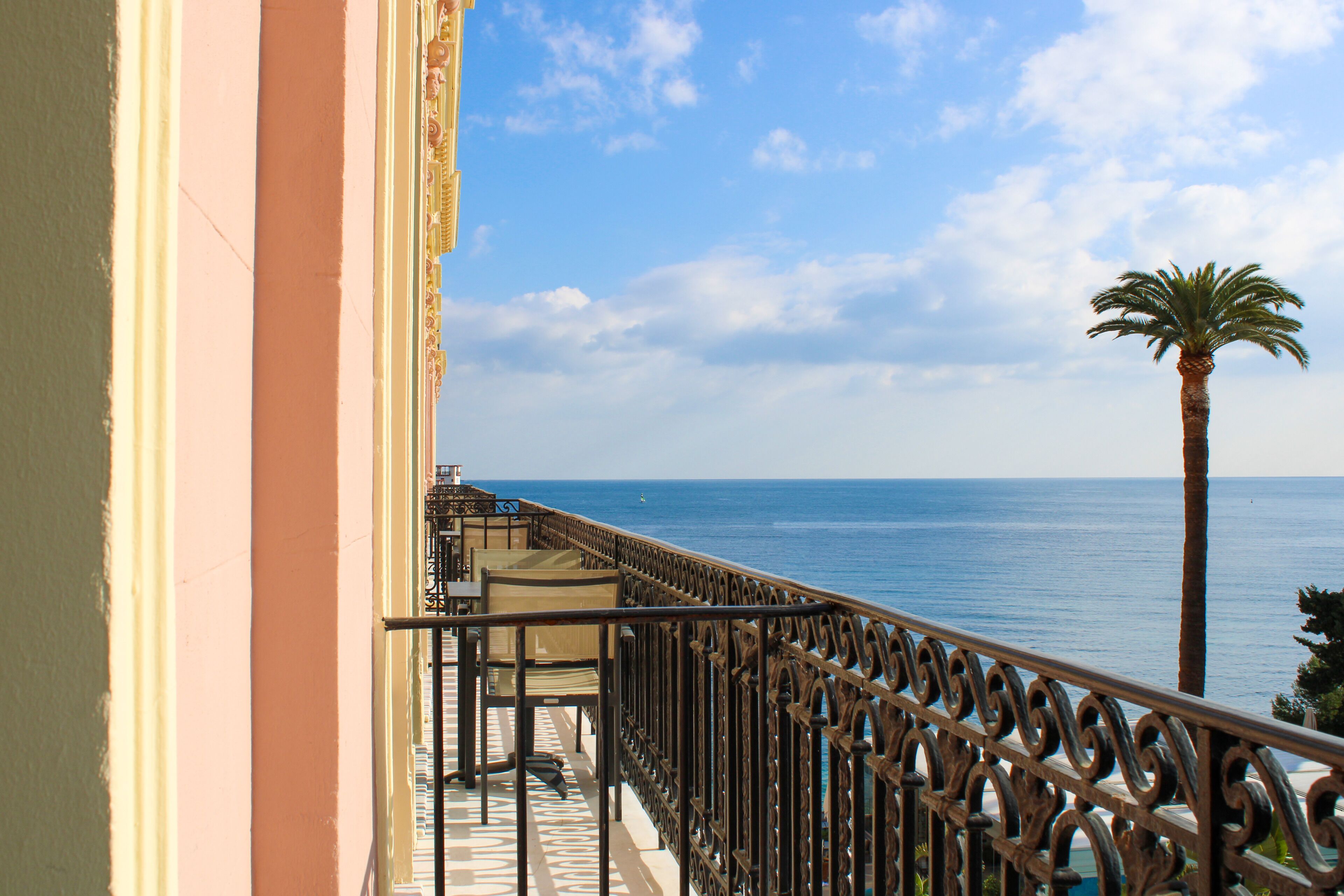 A balcony with a view of the sea, a palm tree, and a building with a pink facade.