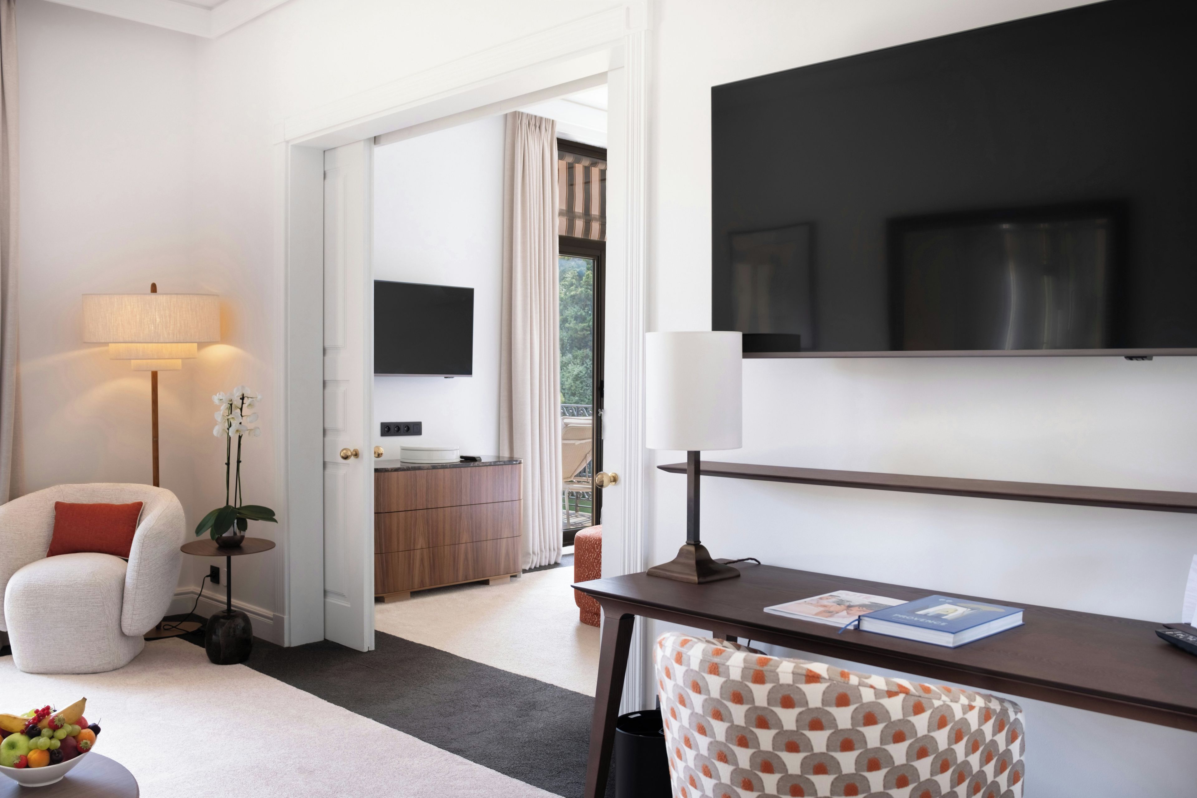 A hotel room with a bed, a desk, a chair, a TV, a lamp, a plant, and a fruit bowl.