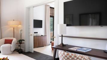 A hotel room with a bed, a desk, a chair, a TV, a lamp, a plant, and a fruit bowl.