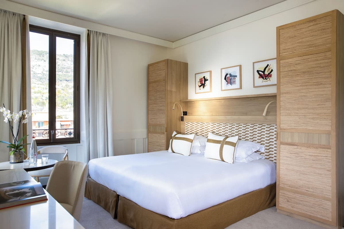 A hotel room with a bed, a desk, a chair, a window with curtains, and wall-mounted artwork.