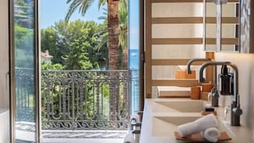 A modern bathroom with a large window, a balcony with ornate metal railings, and a view of palm trees and a body of water.
