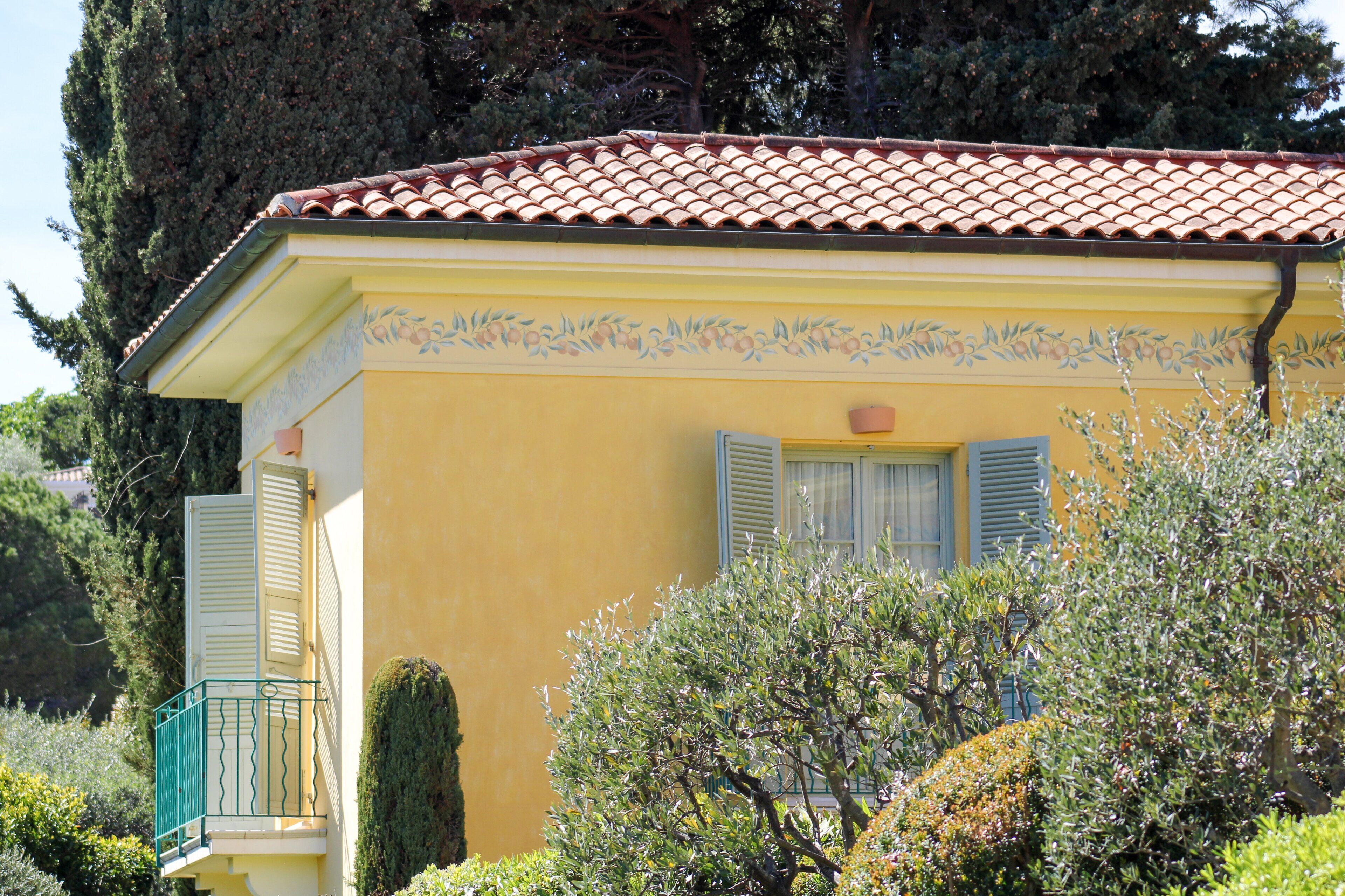 Un bâtiment jaune, doté d’un toit en tuiles et de volets, entouré de verdure.