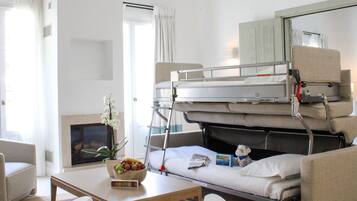 A compact hotel room with a bunk bed, a small table with a bowl of fruit and a teddy bear, a sofa, and a fireplace.