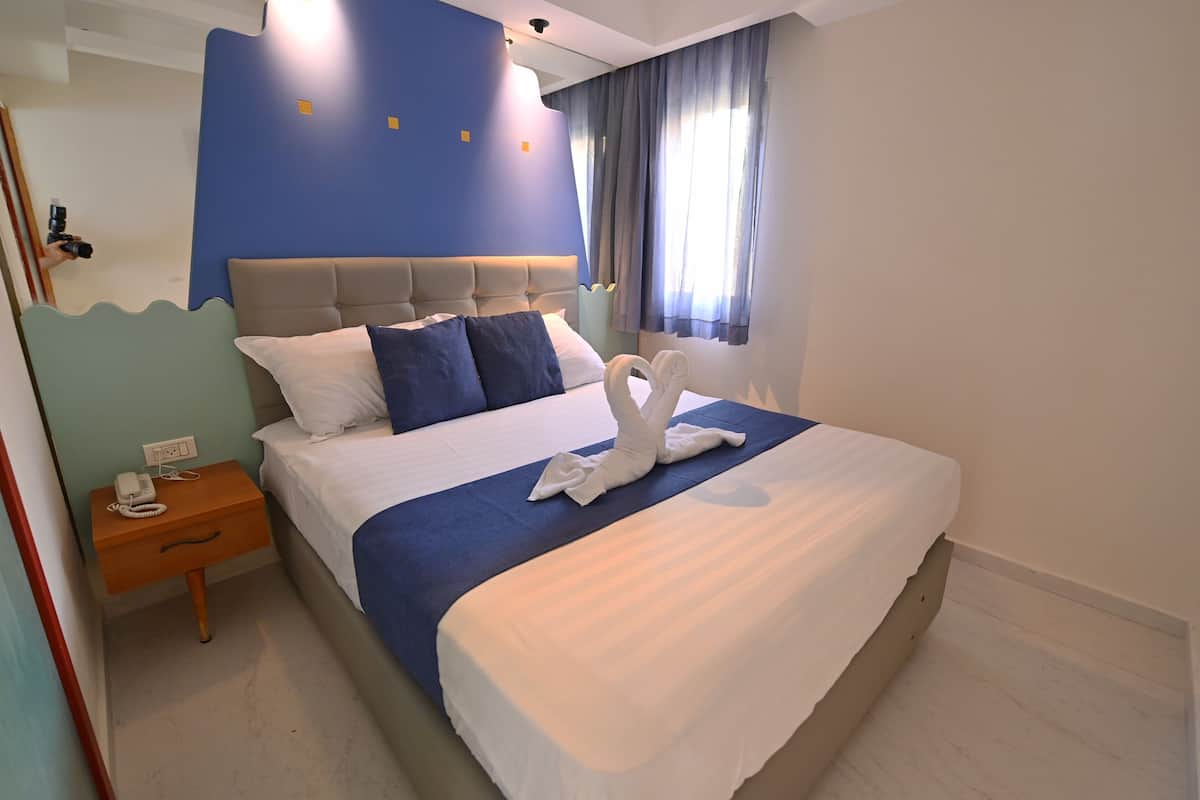 Een keurig opgemaakt bed met een blauwe dekbedovertrek en witte kussens, een nachtkastje met een telefoon, en een raam met gordijnen.