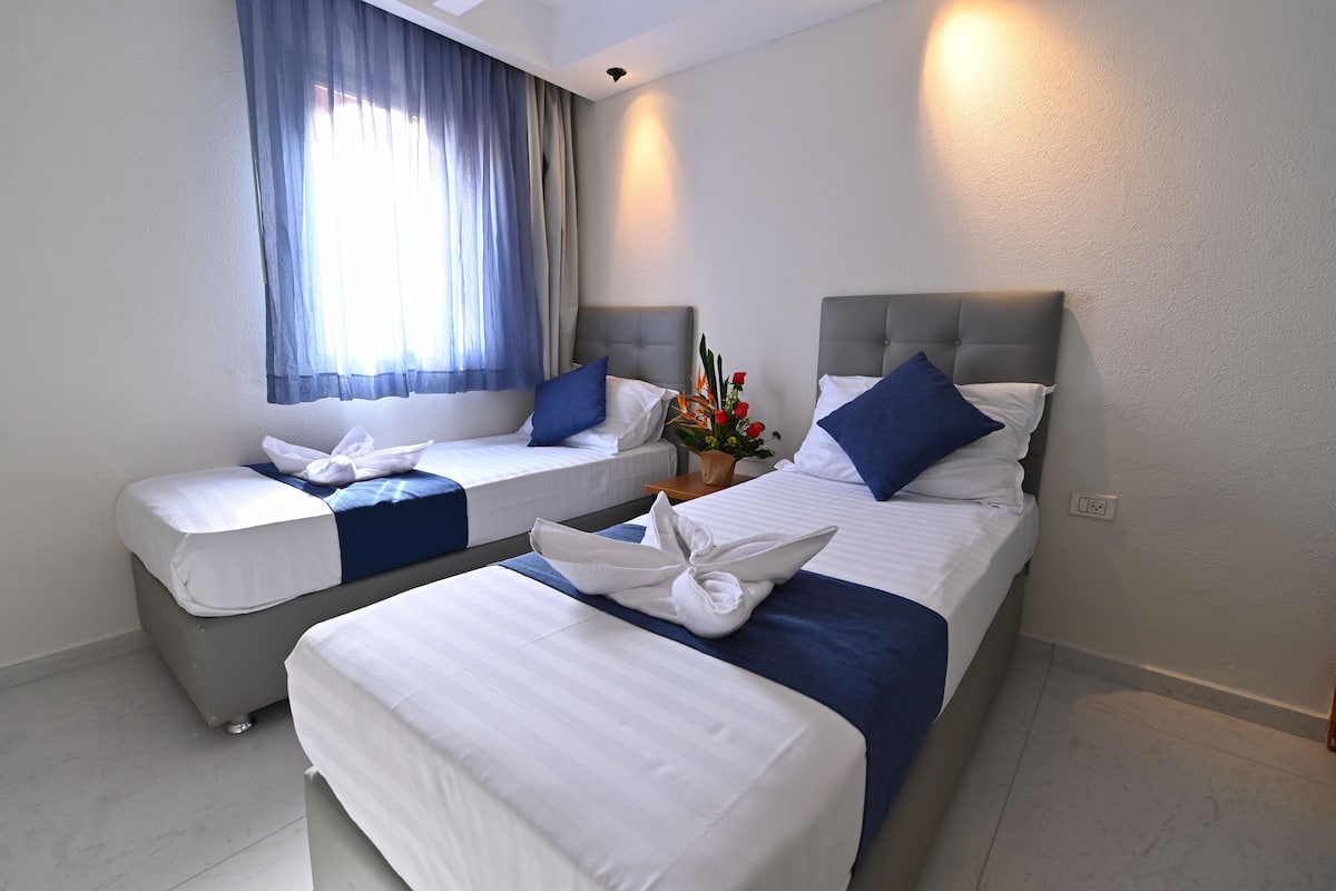 Hotelkamer met twee bedden, elk voorzien van witte lakens en blauwe accenten, een groot raam met dunne gordijnen, en een wandlamp.