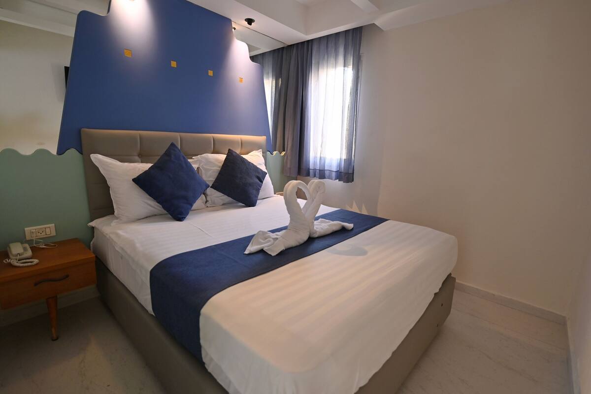 Een netjes opgemaakt bed met witte beddengoed en blauwe kussens, een houten nachtkastje met een telefoon, en een raam met gordijnen.