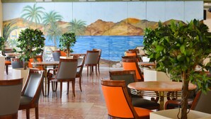 A restaurant with tables and chairs, a mural of a beach scene, and potted plants.