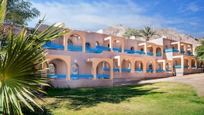 A multi-story building with a series of balconies and arched windows, surrounded by greenery and a mountainous backdrop.