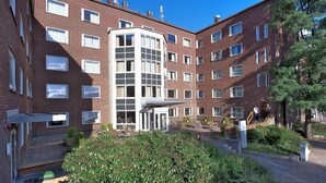 Un immeuble en briques à plusieurs étages, doté d’une entrée centrale, d’un espace extérieur aménagé pour s’asseoir et d’un petit jardin.