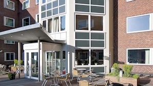 Un hôtel au design moderne, doté d’un espace extérieur couvert avec des tables et des chaises, et d’une façade en briques agrémentée de nombreuses fenêtres.