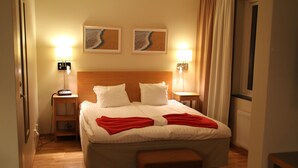 Une chambre d’hôtel avec un grand lit, deux tables de chevet, une petite table, un banc en bois et deux lampes fixées au mur.