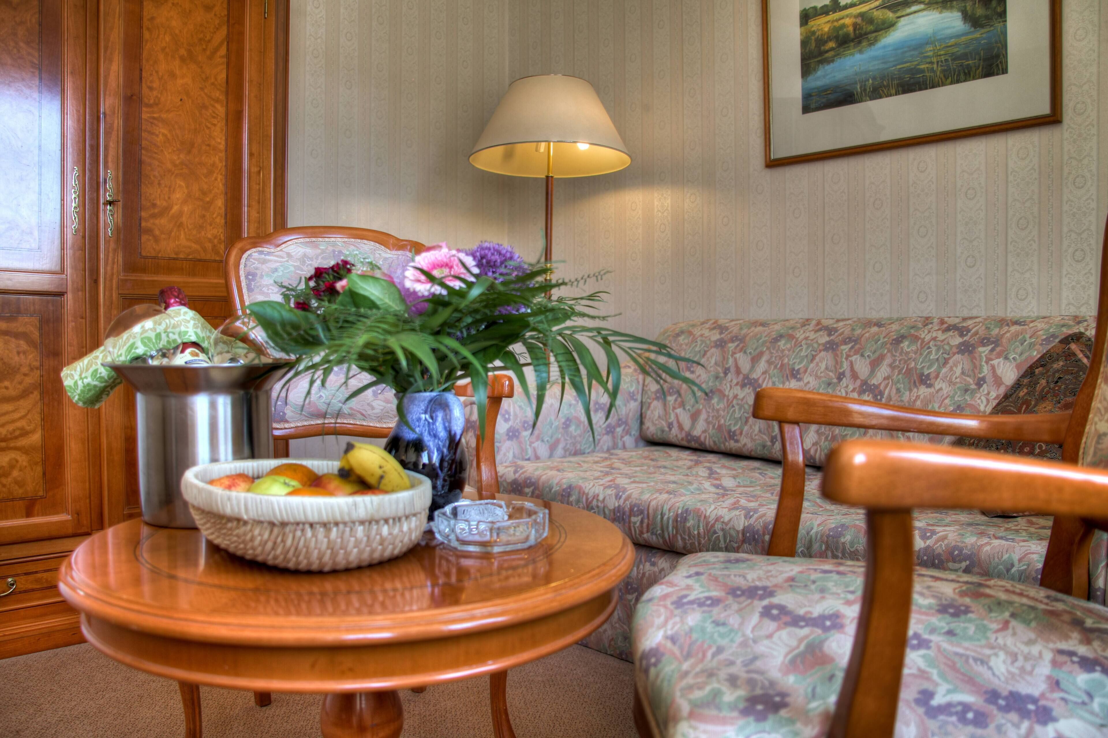 A cozy living room with a wooden cabinet, a sofa, a round table with a fruit bowl, a vase with flowers, and a lamp.