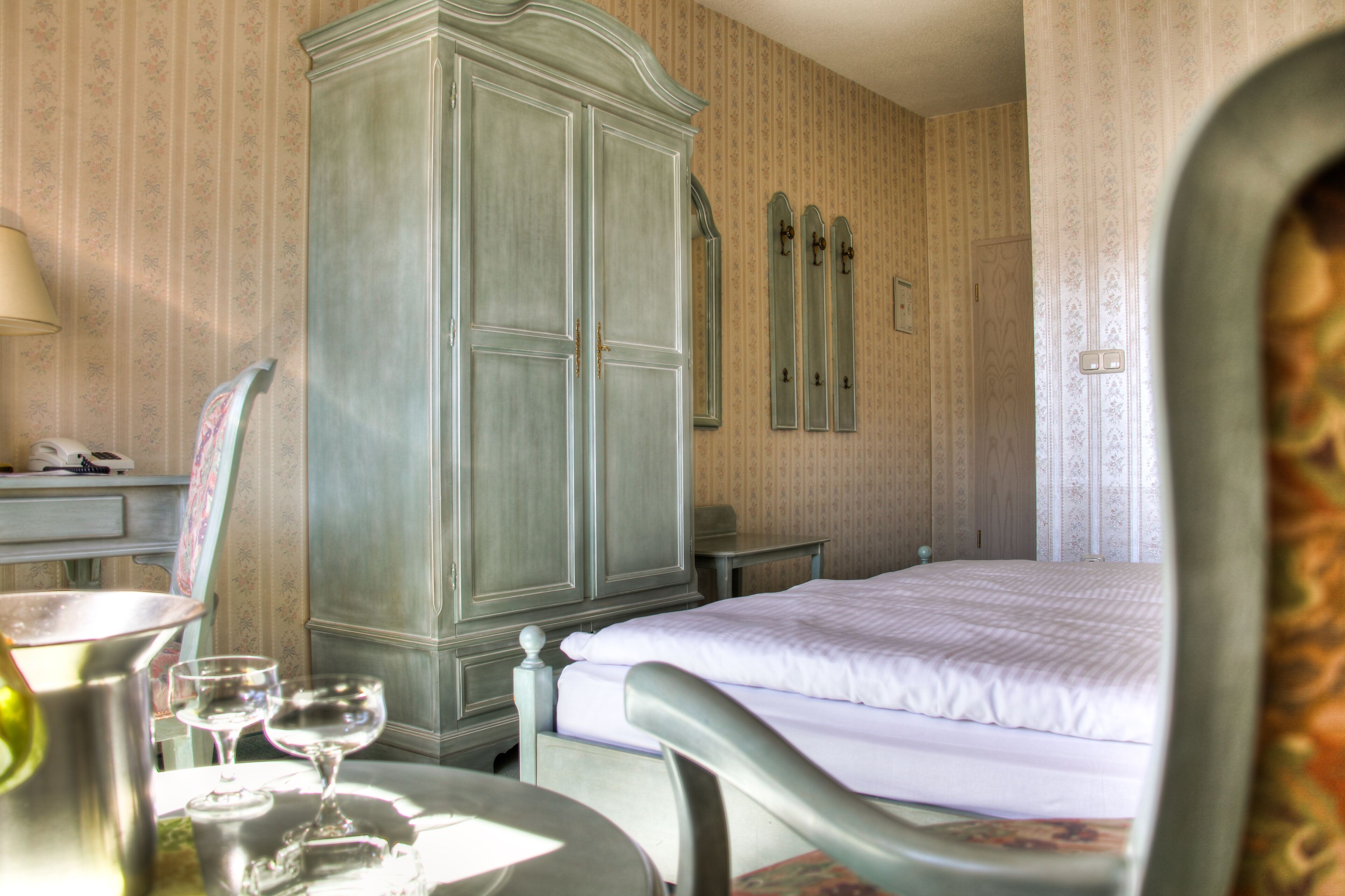 A hotel room with a bed, a wardrobe, a table with glasses, and a lamp.