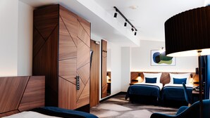 A modern hotel room with a large wardrobe, two beds with blue bedding, a desk with a lamp, and a large abstract painting on the wall. - Hotel Ascot Premium (Kraków)