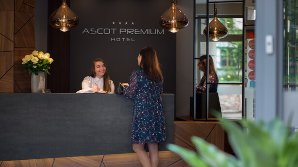A reception desk at Ascott Premium Hotel with a modern design, a black wall with the hotel name, and a large window offering a view of the outdoors. - Hotel Ascot Premium (Kraków)