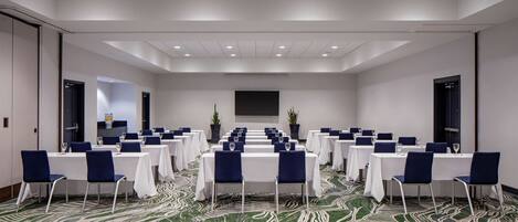 Un salón de conferencias con filas de mesas y sillas, una pantalla grande y una alfombra estampada.