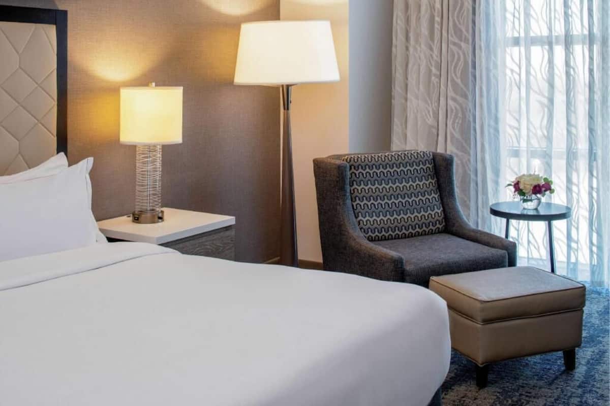 Hotellihuone, jossa on sänky, yöpöytä lampun kanssa, kuviollinen nojatuoli, pieni pöytä maljakolla ja näkymä ulos.