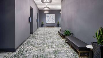 A hallway with a patterned carpet, benches, potted plants, and a large abstract painting on the wall.