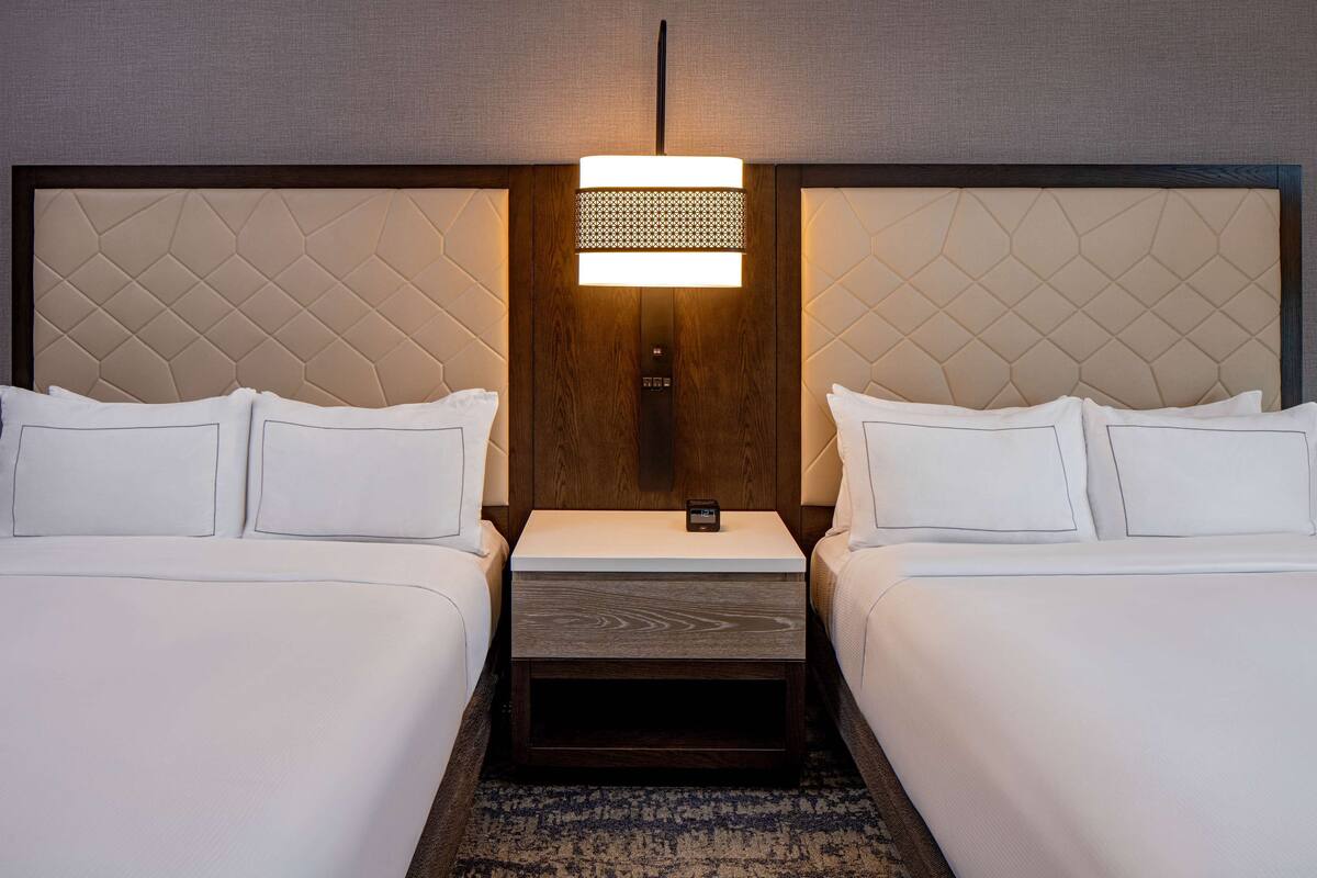 Hotellihuone, jossa on kaksi sänkyä, yöpöytä ja seinään kiinnitetty valaisin.