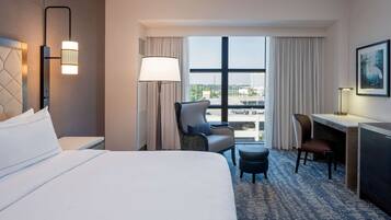A hotel room with a bed, a desk, a chair, and a view of the city through large windows.