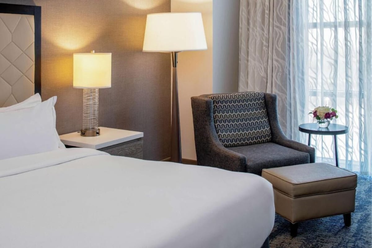 Un cuarto de hotel con cama, una mesita de noche con lámpara, un sillón estampado, una pequeña mesa con un florero, y vista al exterior.