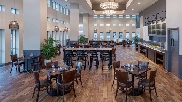 A spacious dining area with multiple tables and chairs, large windows, and a central island with seating. - Hilton Shreveport (Shreveport)