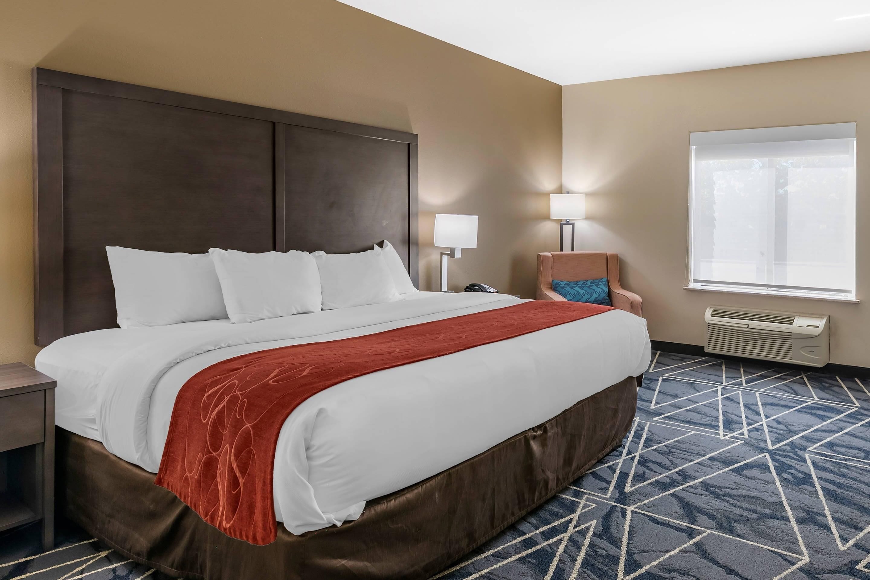 A hotel room with a large bed, a nightstand, a chair, a window with blinds, and an air conditioning unit.