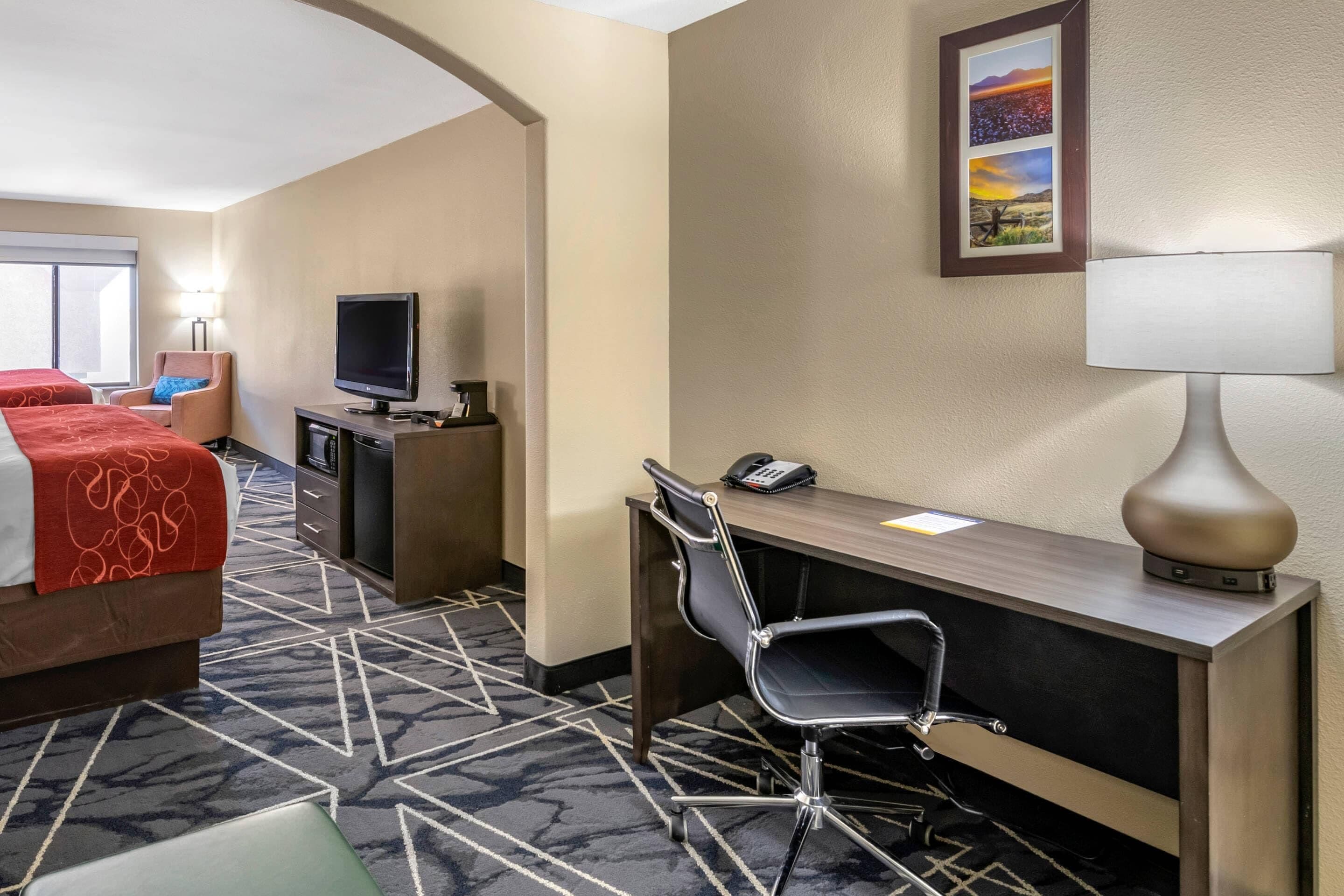 Habitación de hotel con cama, escritorio, silla, televisión, lámpara y una obra de arte en la pared.