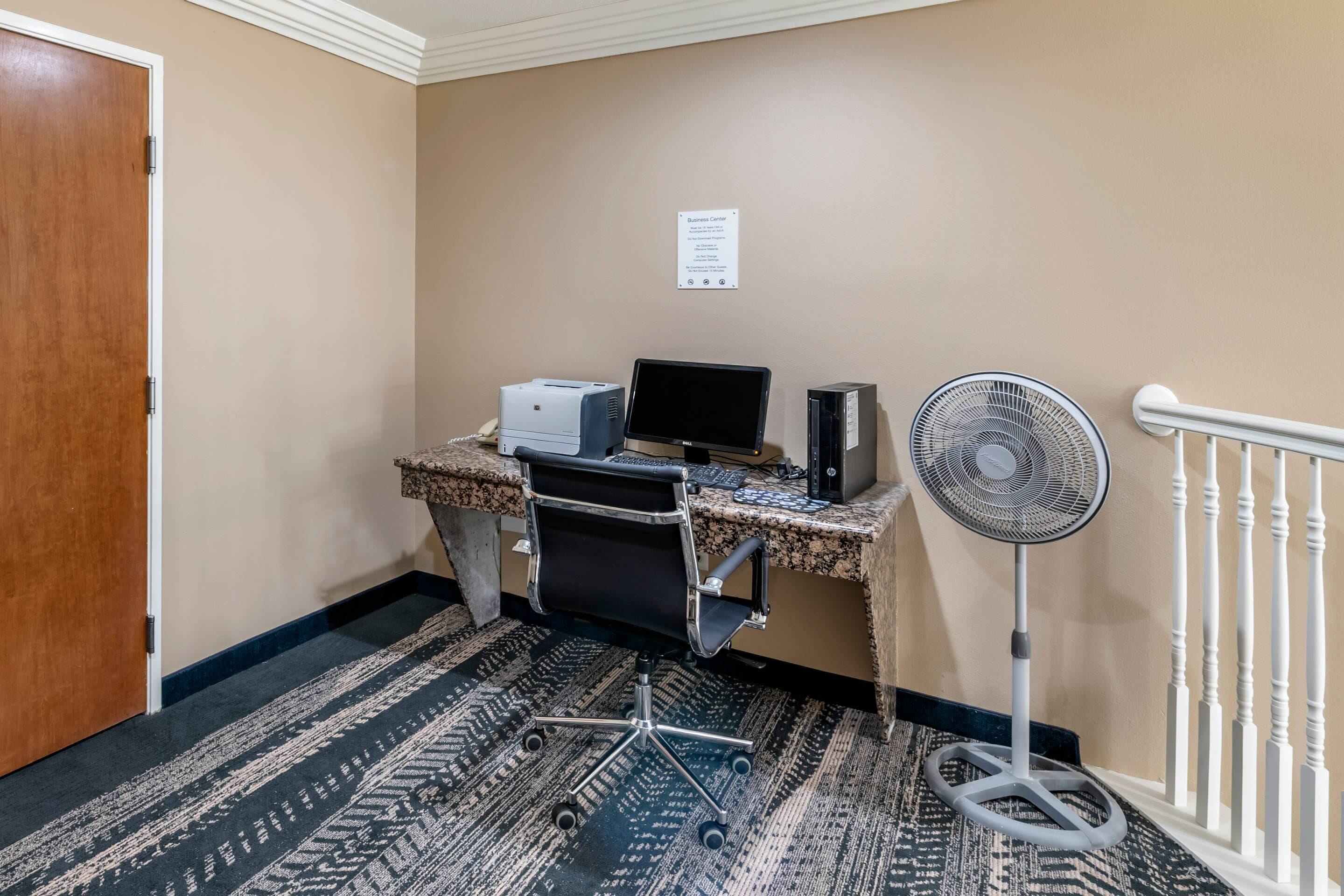 A hotel room with a desk, computer, printer, and a standing fan.