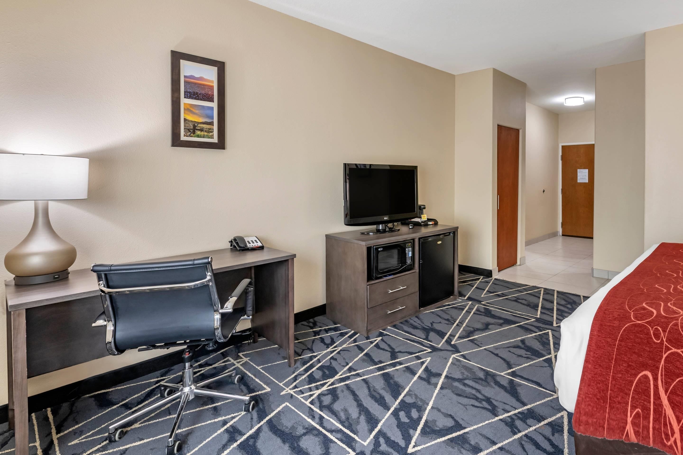 A hotel room with a desk, chair, TV, microwave, and a bed with a red blanket.