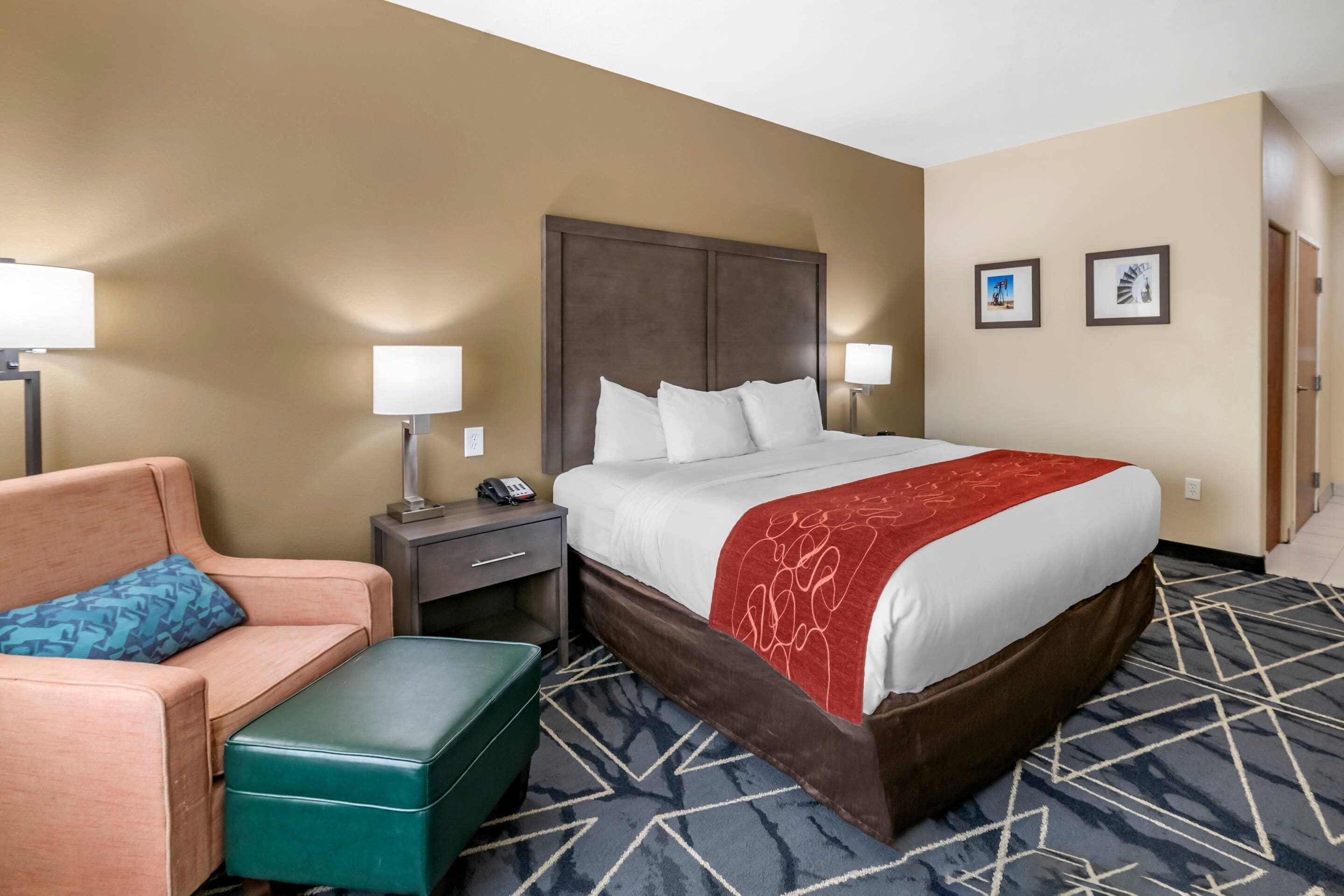 A hotel room with a large bed, a green ottoman, a sofa, a nightstand, and two wall-mounted lamps.