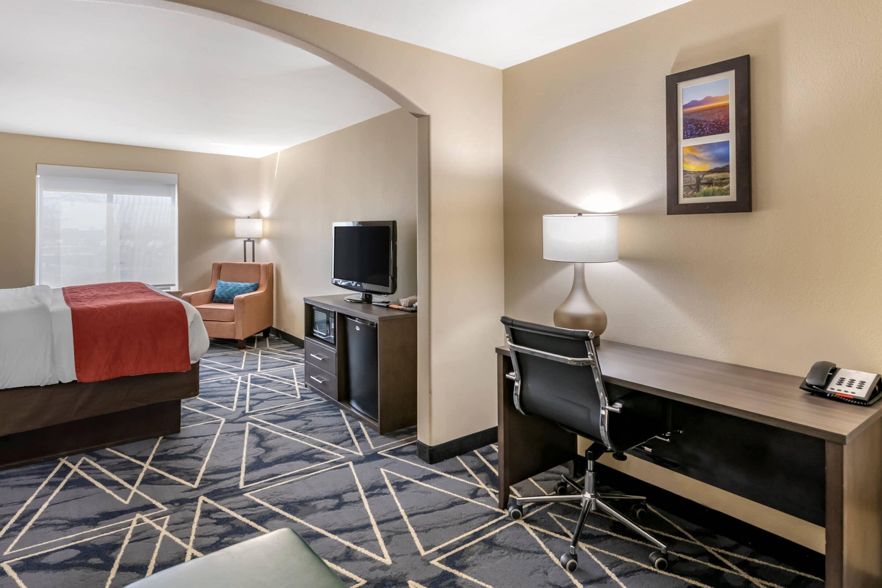 A hotel room with a bed, desk, chair, TV, and a painting on the wall.