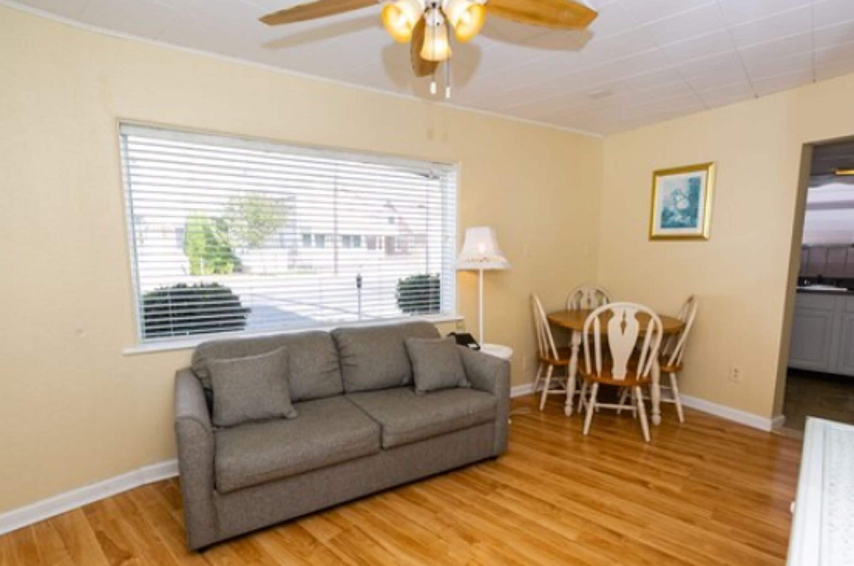 A living room with a sofa, dining table, chairs, a ceiling fan, and a window with blinds.
