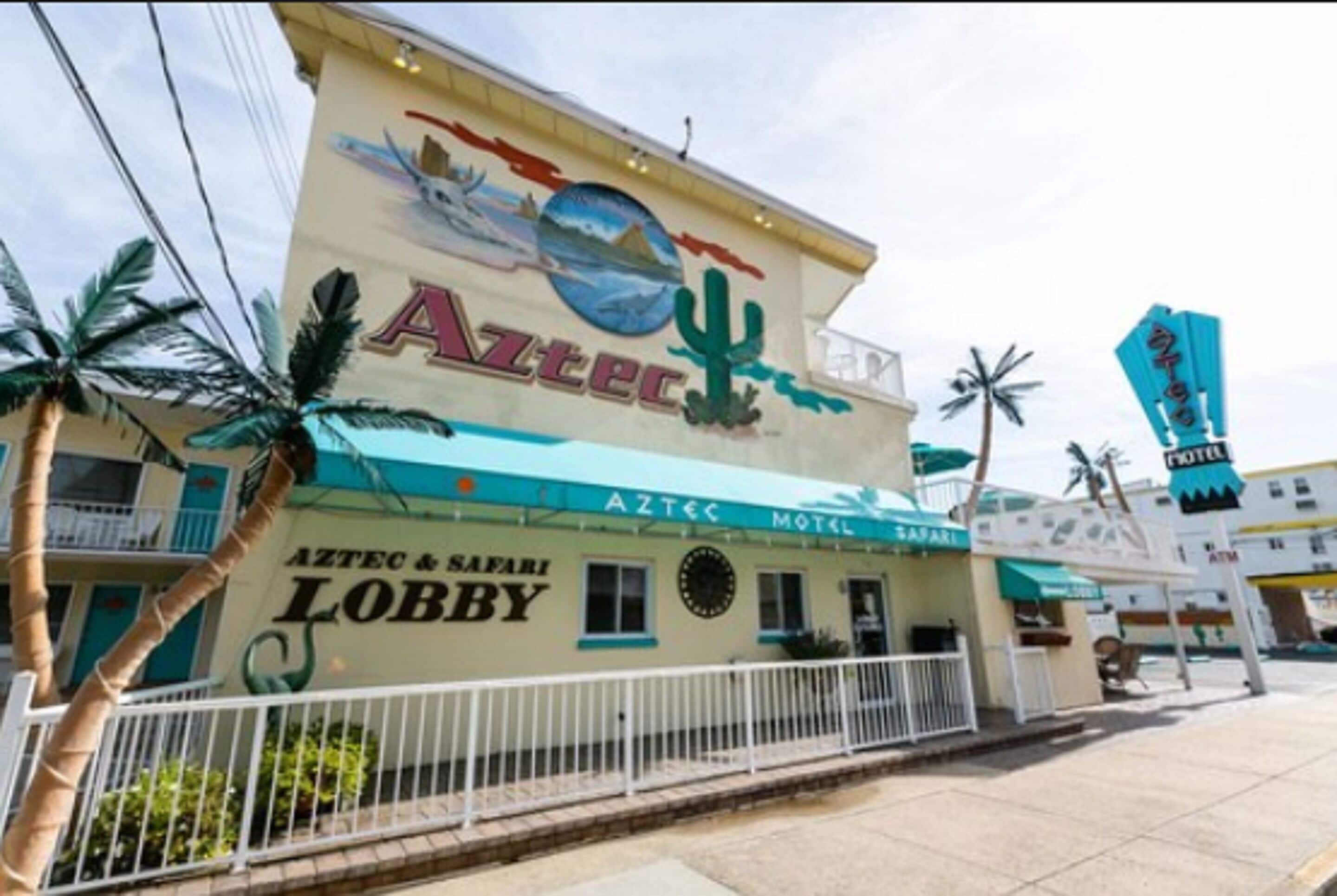 The exterior of the Aztec Motel, featuring a prominent sign with a desert landscape and cacti, and the name 'AZTEC MOTEL' displayed on the facade.