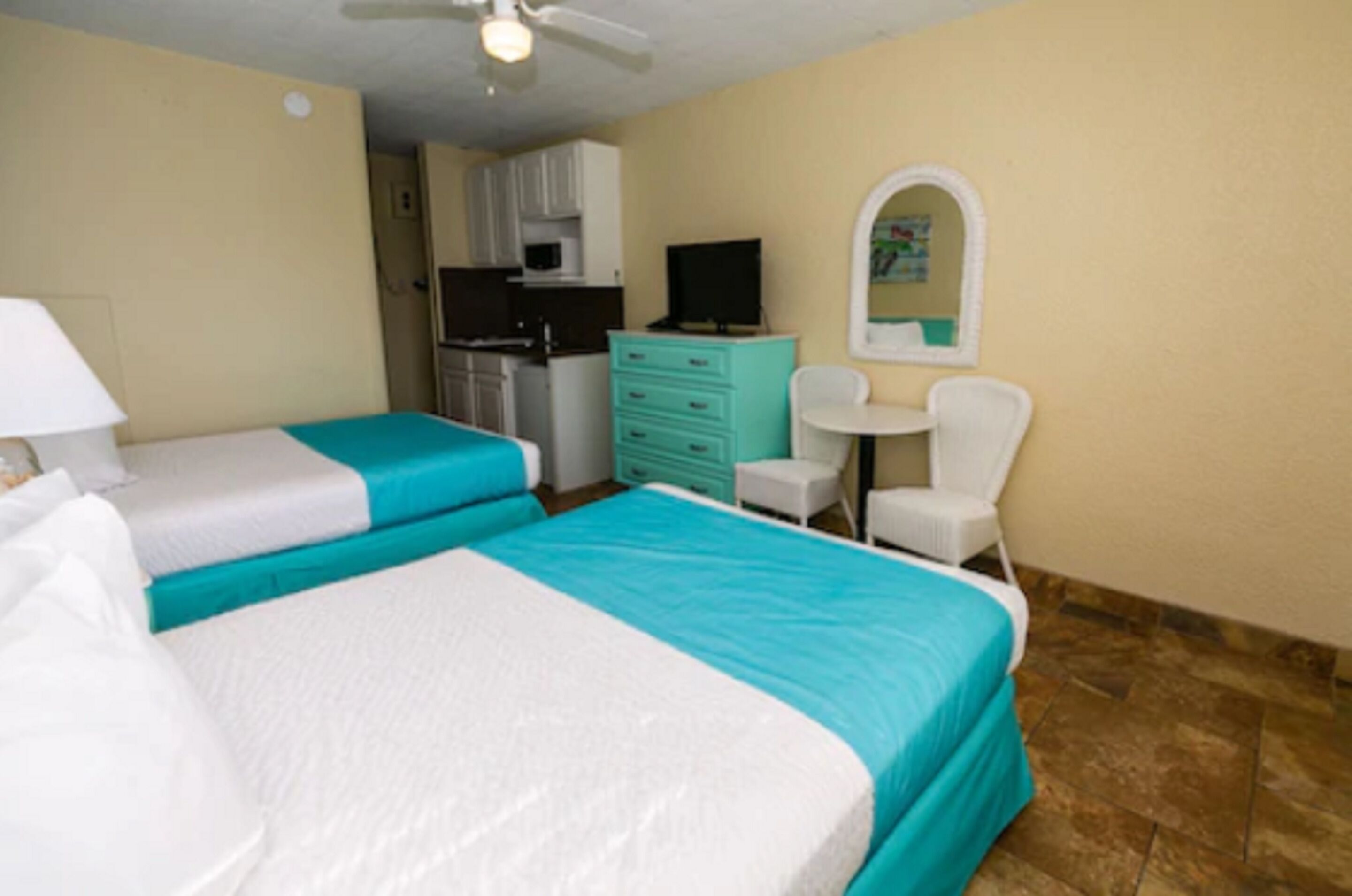 A hotel room with two beds, a kitchenette, a small table with a chair, and a mirror on the wall.