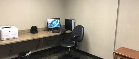 A corner office with a desktop computer, a printer, a trash bin, and a small wooden table.