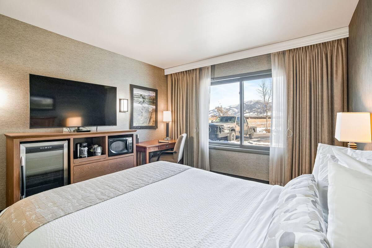 A hotel room with a bed, a TV, a desk with a chair, a minibar, and a window with curtains.