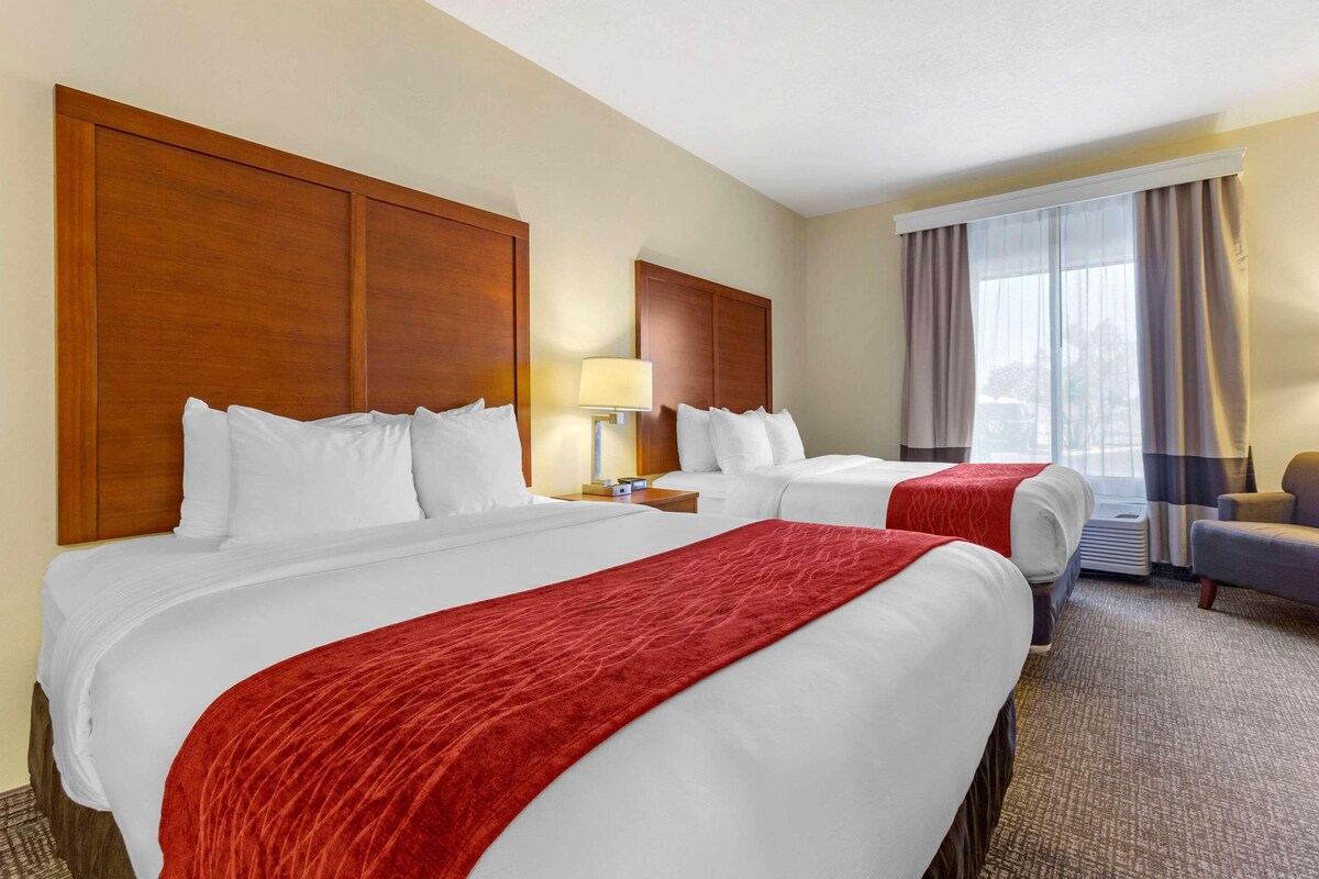 A hotel room with two beds, a desk, a chair, and a large window with curtains.