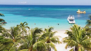 A beach with clear turquoise water, palm trees, and boats. - Henry Morgan Resort - All Inclusive (Roatan)
