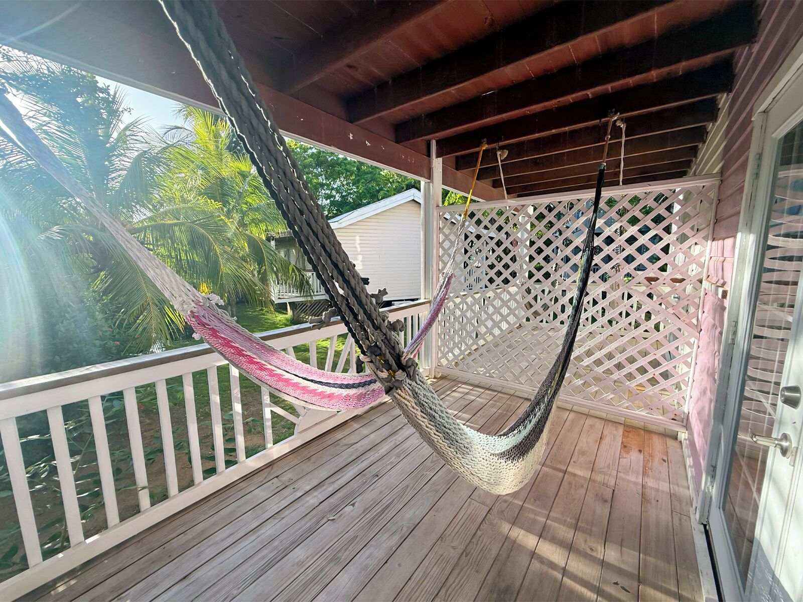 A hammock on a wooden deck with a lattice fence and a view of a house and palm trees.