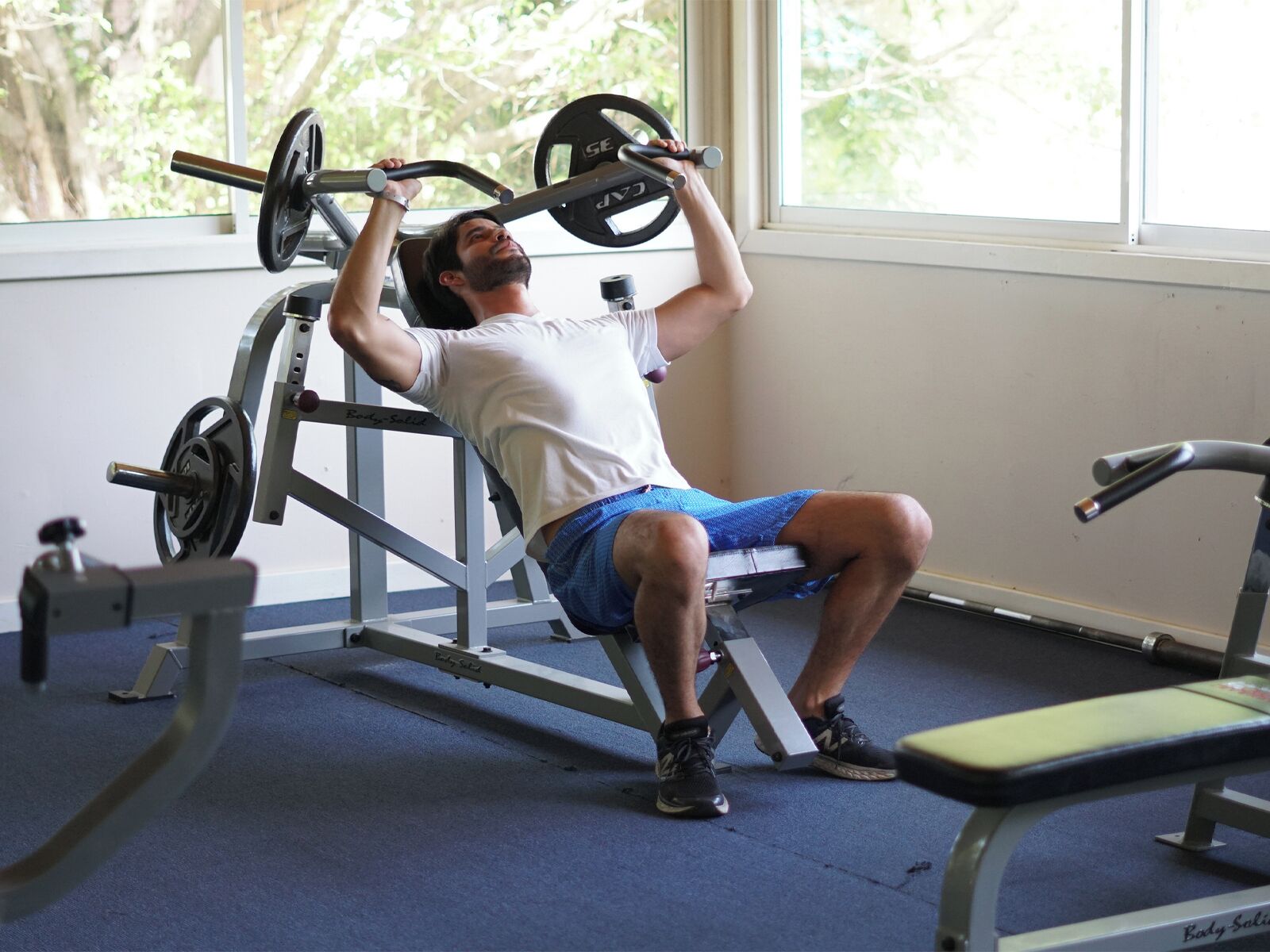 En person som använder en bänkpressmaskin i ett gym, med ett fönster synligt i bakgrunden.