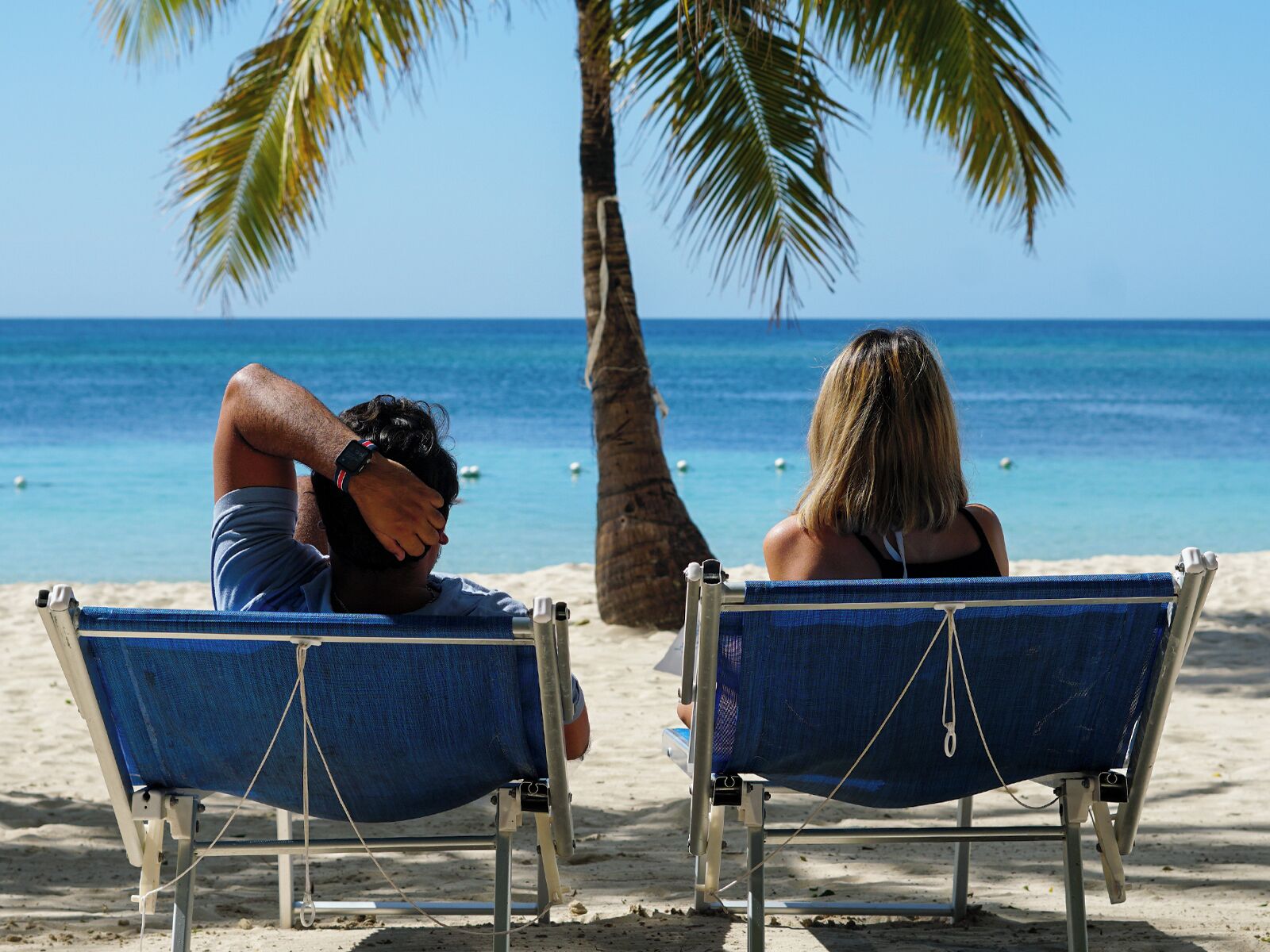 Two beach chairs facing the ocean with a palm tree and clear blue water in the background.