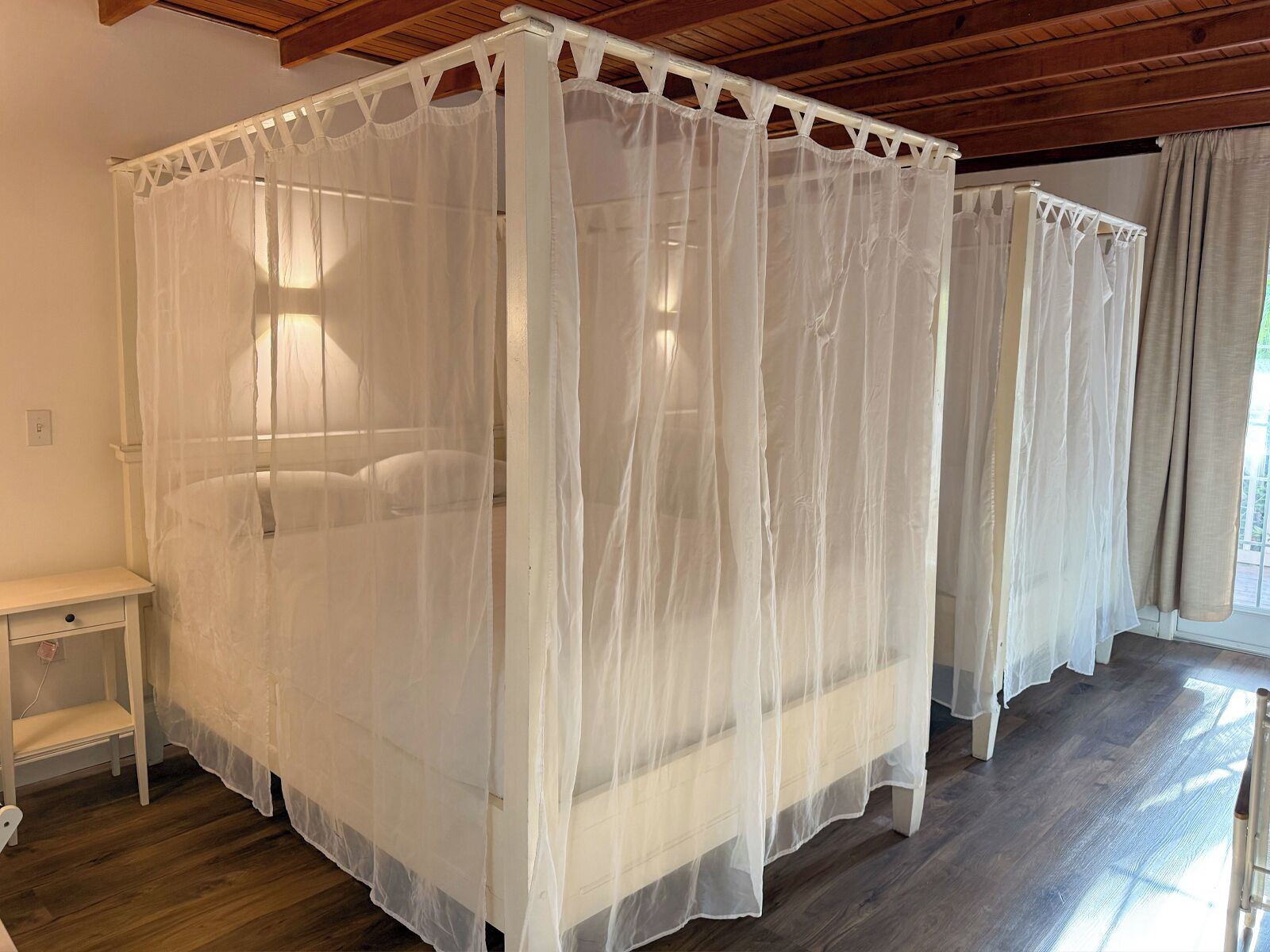 A four-poster canopy bed with a white bedspread and matching canopy, a wooden nightstand, and a window with curtains.